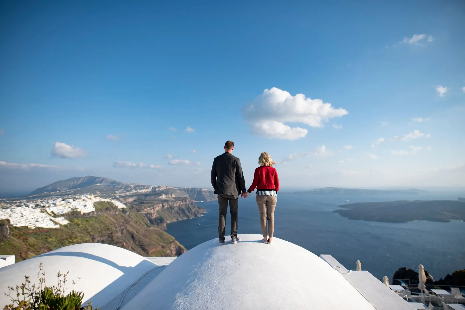 Ege kıyısında, beyaz badanalı bir kubbenin üzerinde el ele duran çiftin denize ve kıyı şeridine bakan görüntüsü.