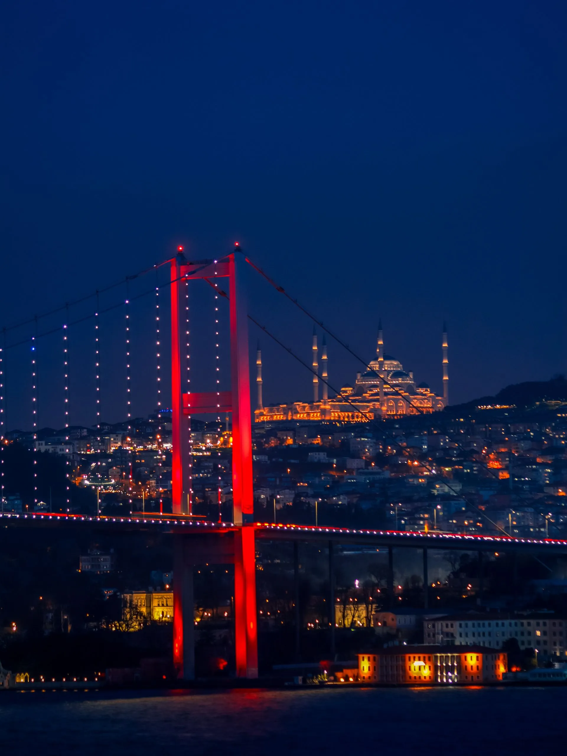 Gece vakti kırmızı ışıkla aydınlatılmış Boğaziçi Köprüsü ve arka planda ışıklandırılmış bir cami siluetı.