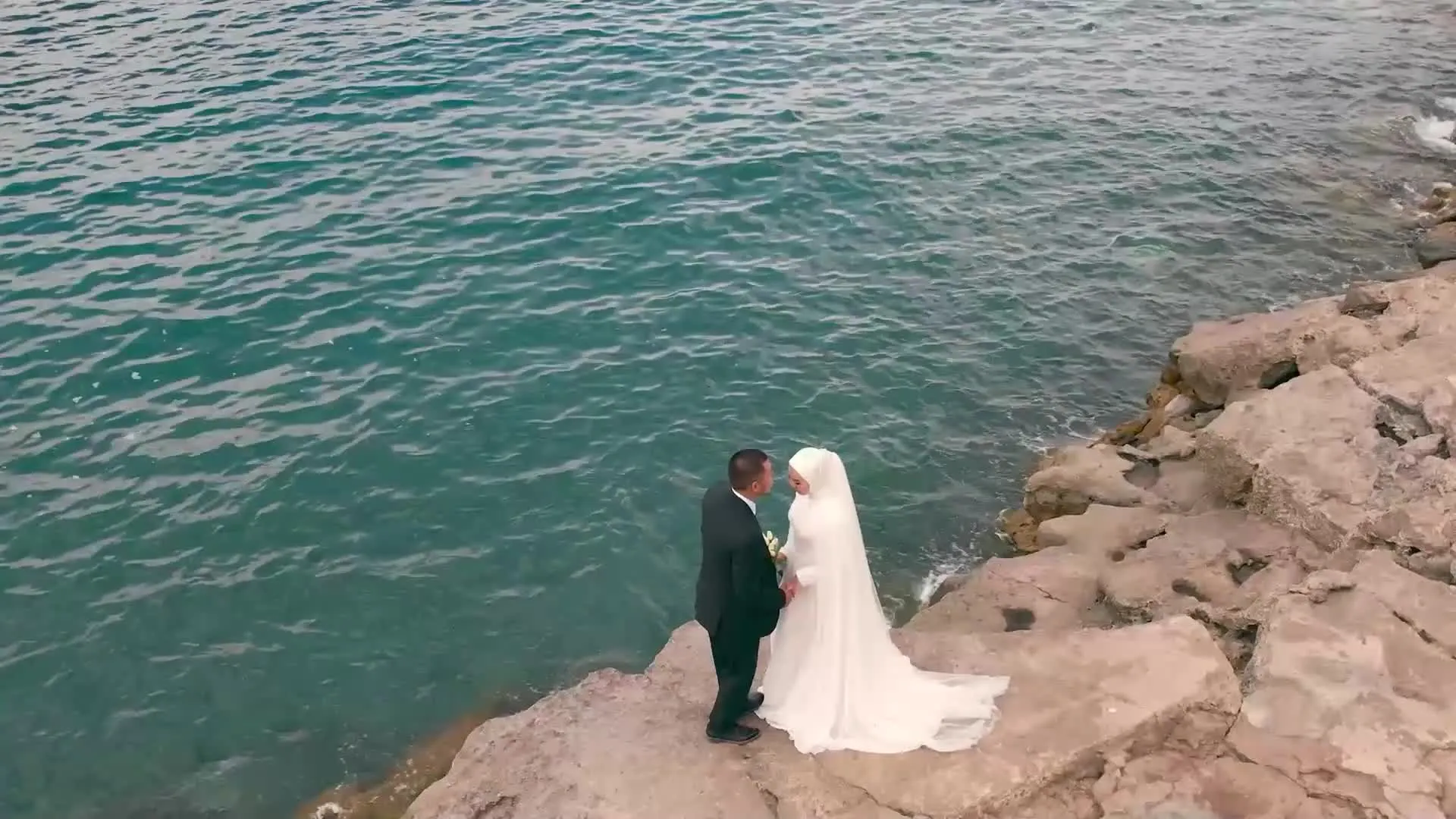 Kayalık bir sahil şeridinde duran gelin ve damadın, denize bakarken drone ile çekilmiş görüntüsü.