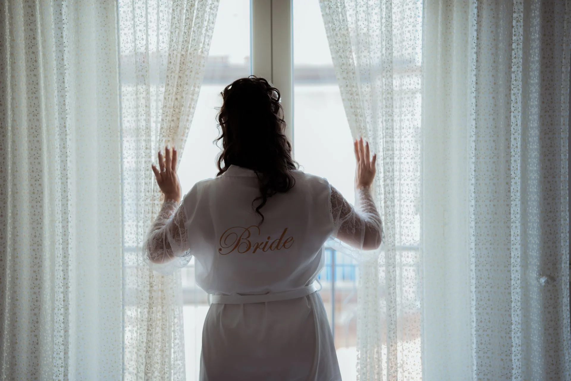 Sırtında 'Bride' yazılı saten sabahlık giyen gelinin, pencereden dışarı bakarken arkadan çekilmiş sessiz anı.